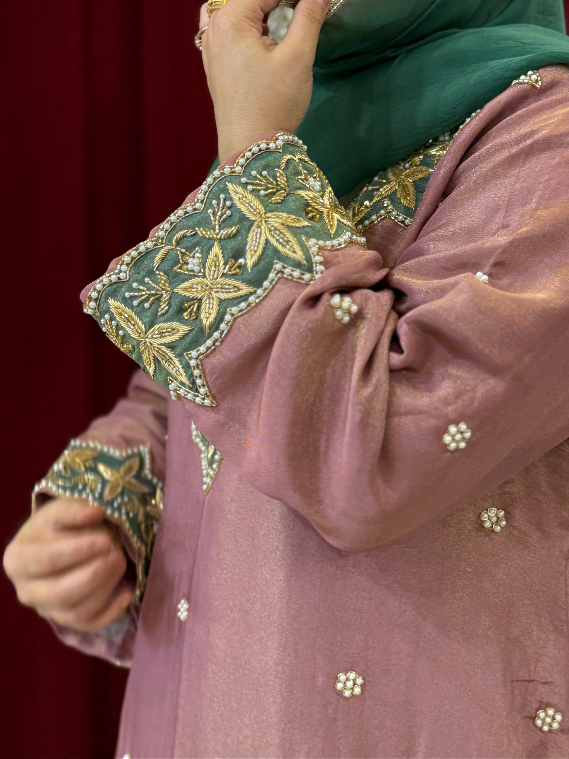Person wearing a traditional outfit with green hijab and embroidered sleeves against a red curtain background